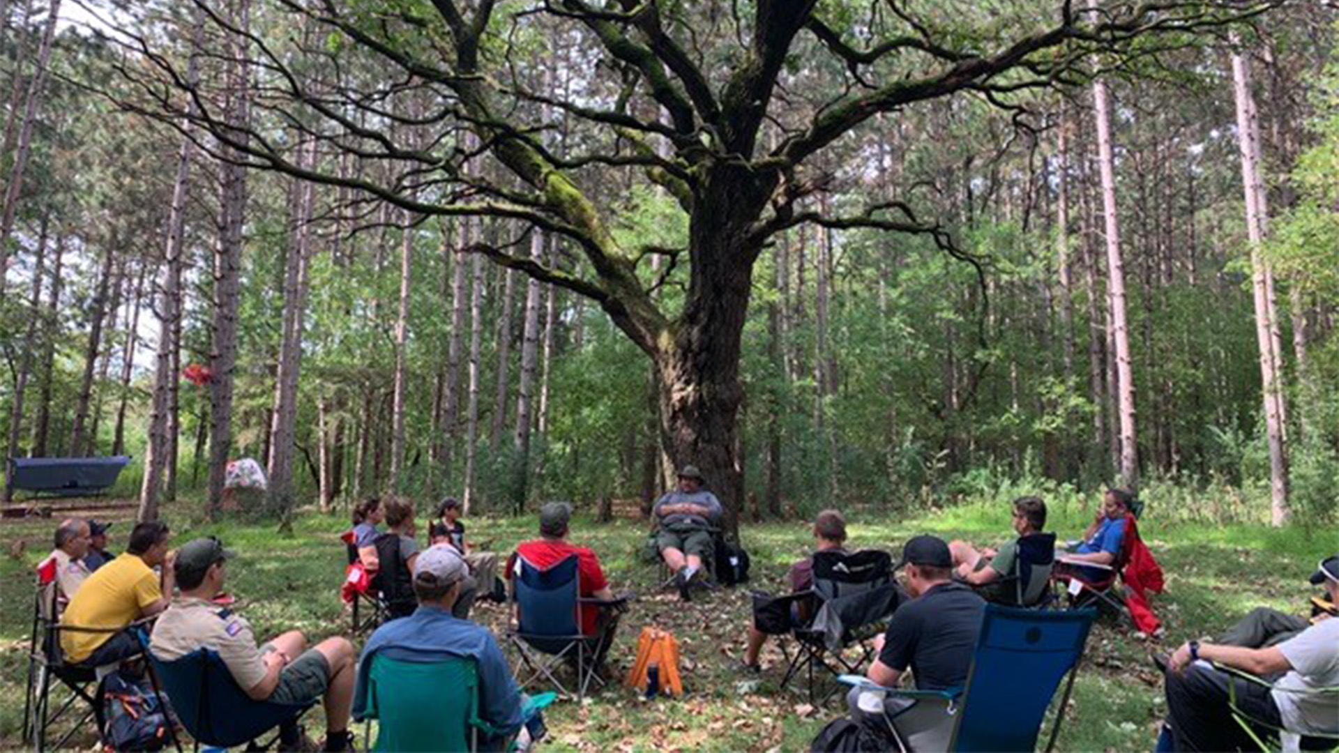 A group of adult leaders sitting in camping chairs outdoors are watching another leader as they lead a discussion. 