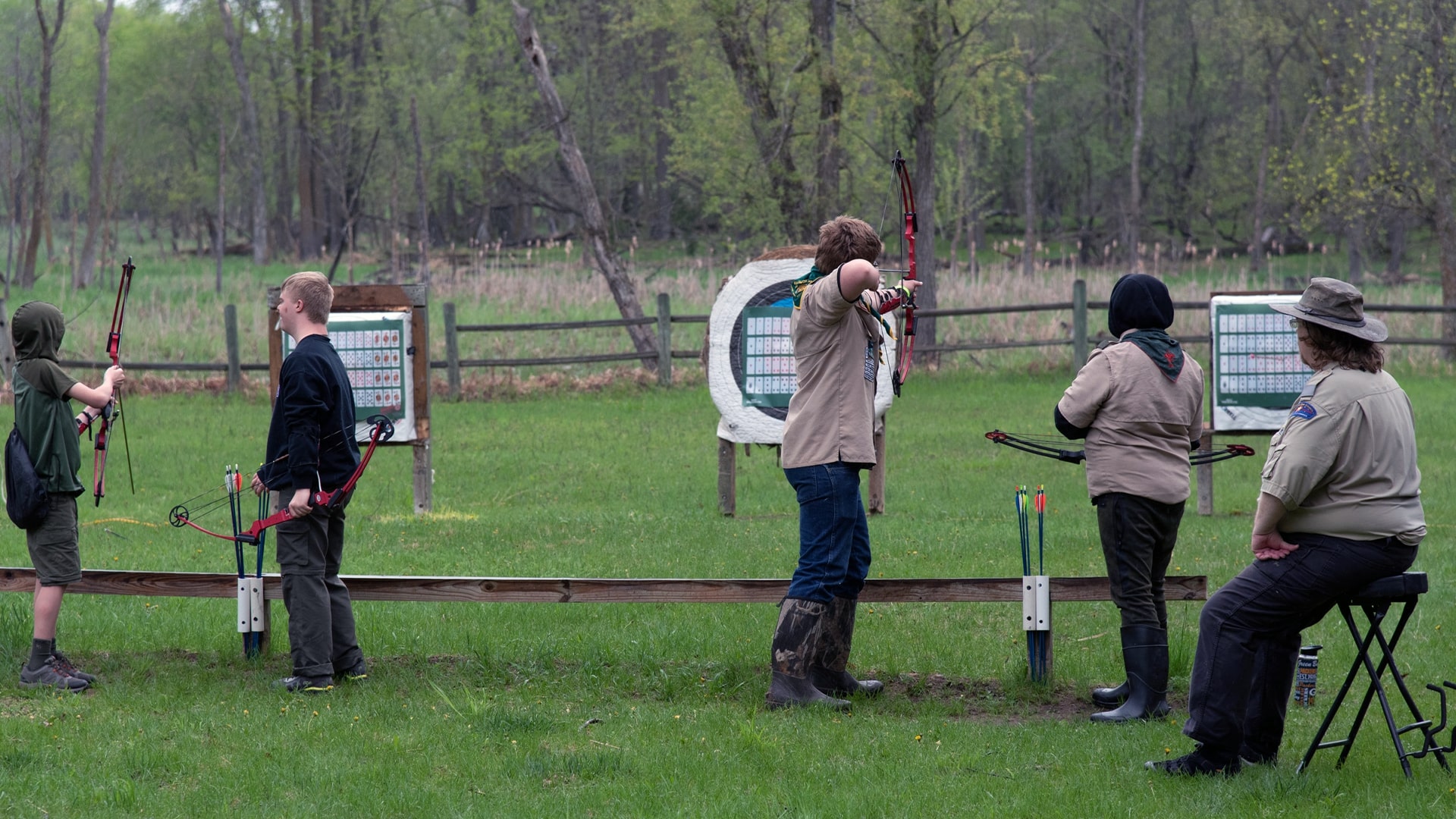 Four Scouts, 2 wearing Field (Class A) uniforms and two in casual clothes, are shooting arrows at the targets down-range while a staff member watches over them.