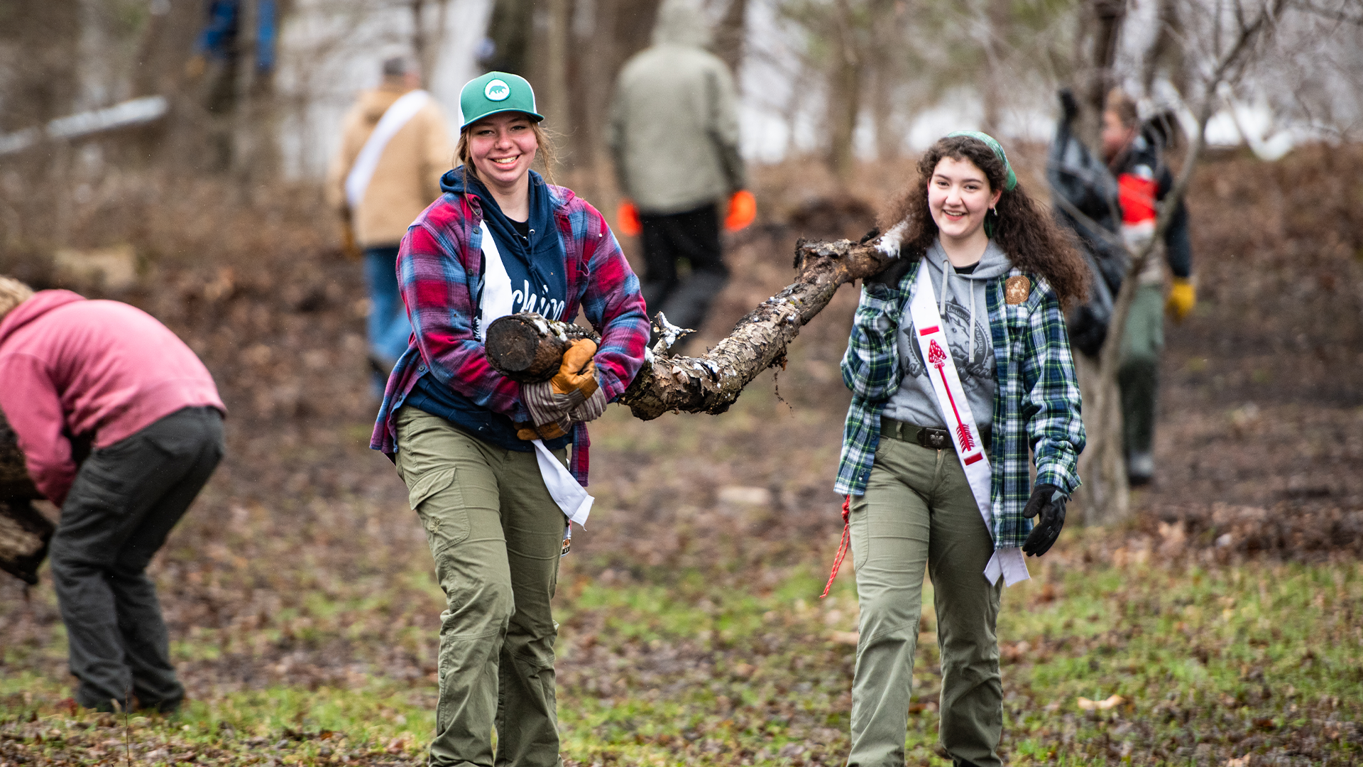 Two female Scouts wearing OA sashes are smiling while carrying a large tree branch as part of a cleanup project. Four other people are seen in the background also working on cleaning up.