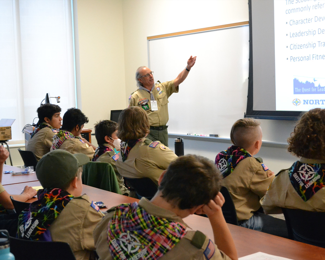 A large group of Scouts in uniform are sitting at desks in a classroom while an adult leader is pointing to a lesson slide outside of the frame