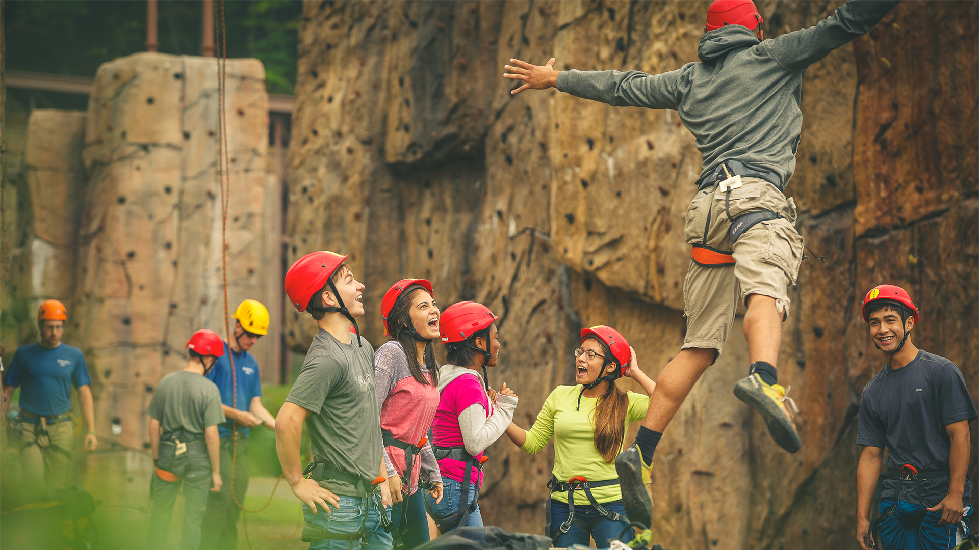 A group of Venturing Scouts belay or watch and cheer for a Venturing Scout climbing an outdoor rock wall. Additional groups of Venturing Scouts can be seen in the background also climbing or belaying and cheering their fellow Scouts up other walls.