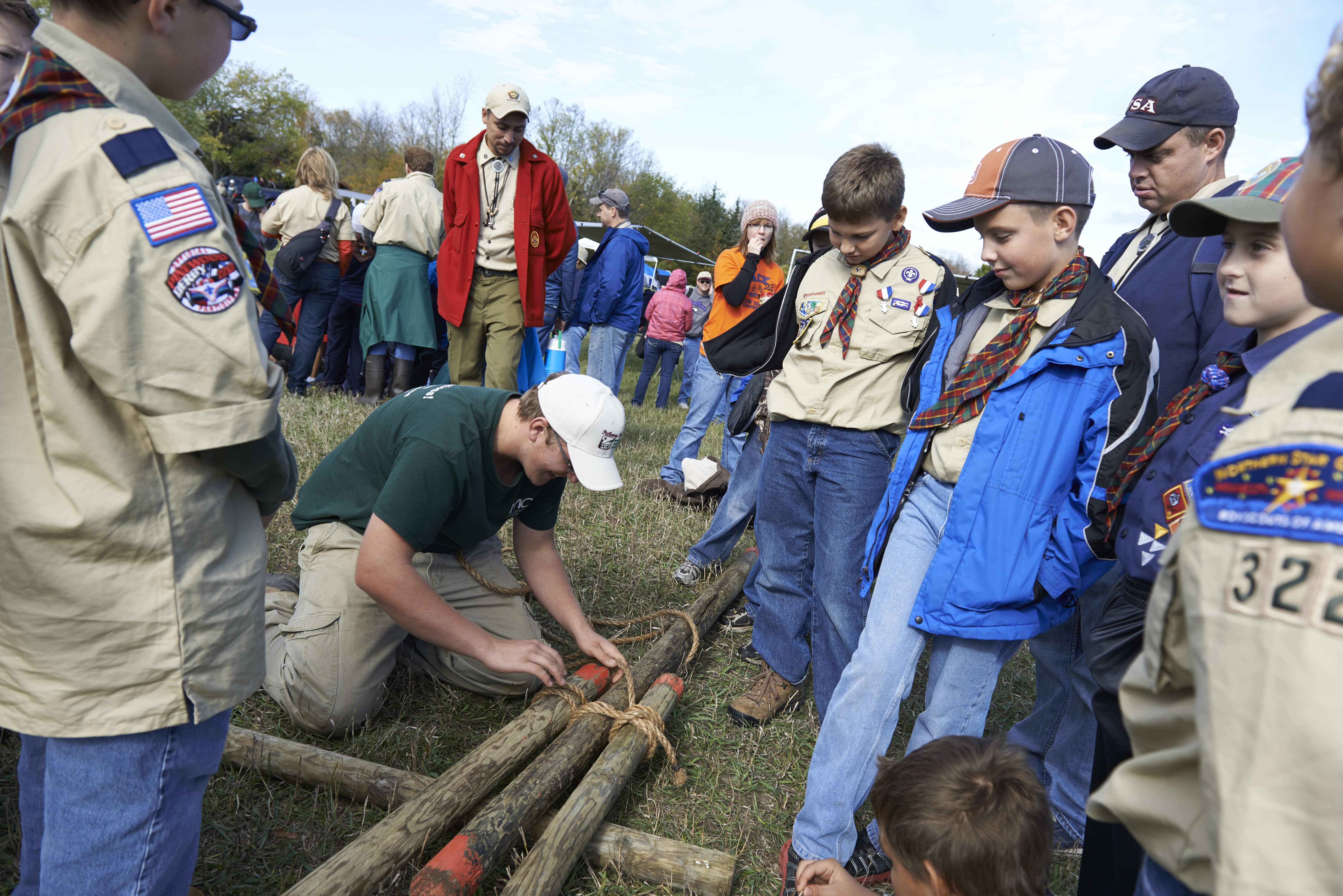 A group of Scouts watching a program counselor tie logs together.