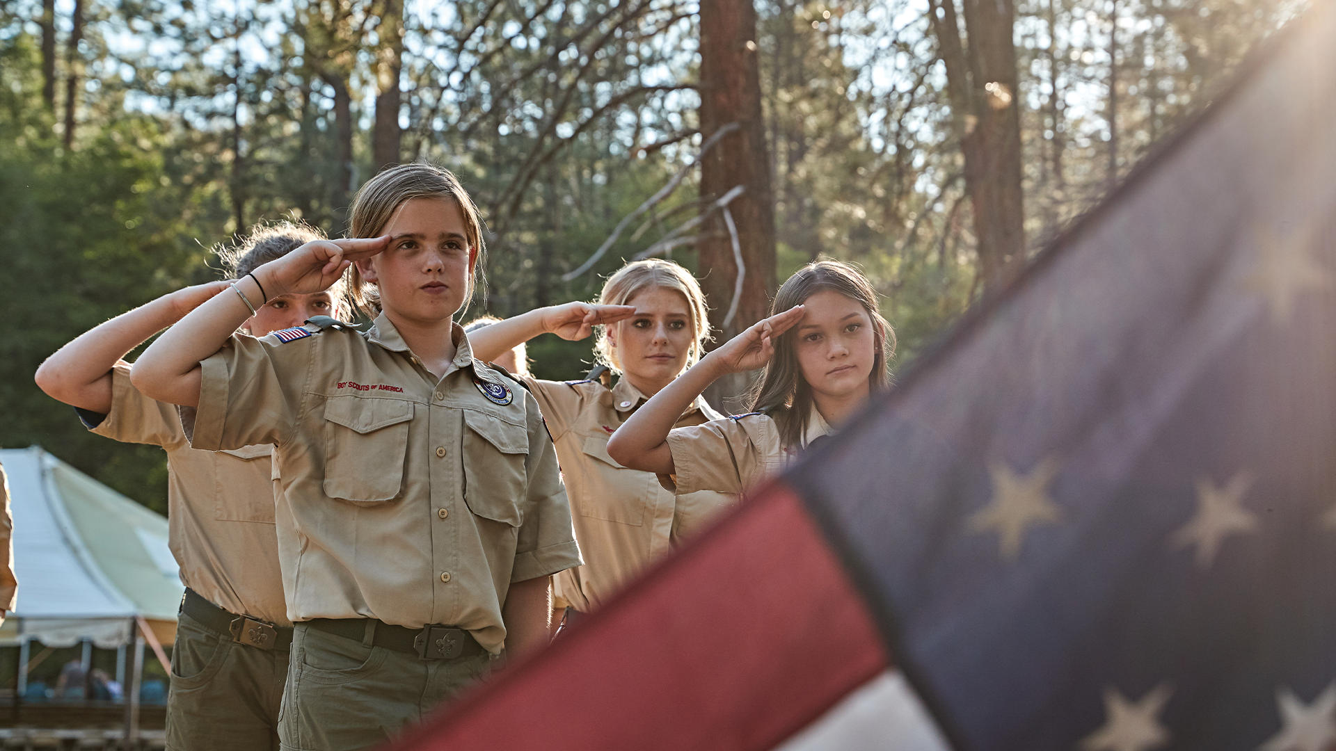 4 female Scouts in a campsite in the woods are saluting the American flag
