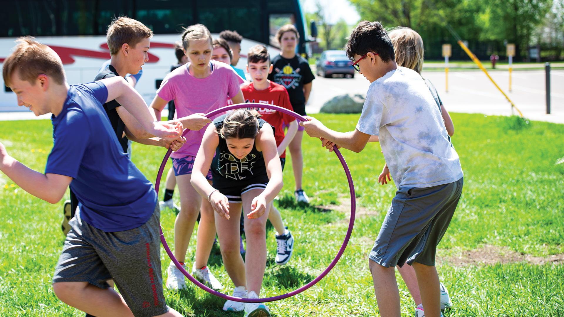 A group of youth playing a game, all looking excited and engaged in playing. One is running away of completing their part, two are hold a hula hoop sideways, and a line has forms with individuals attempting to run through the hula hoop.