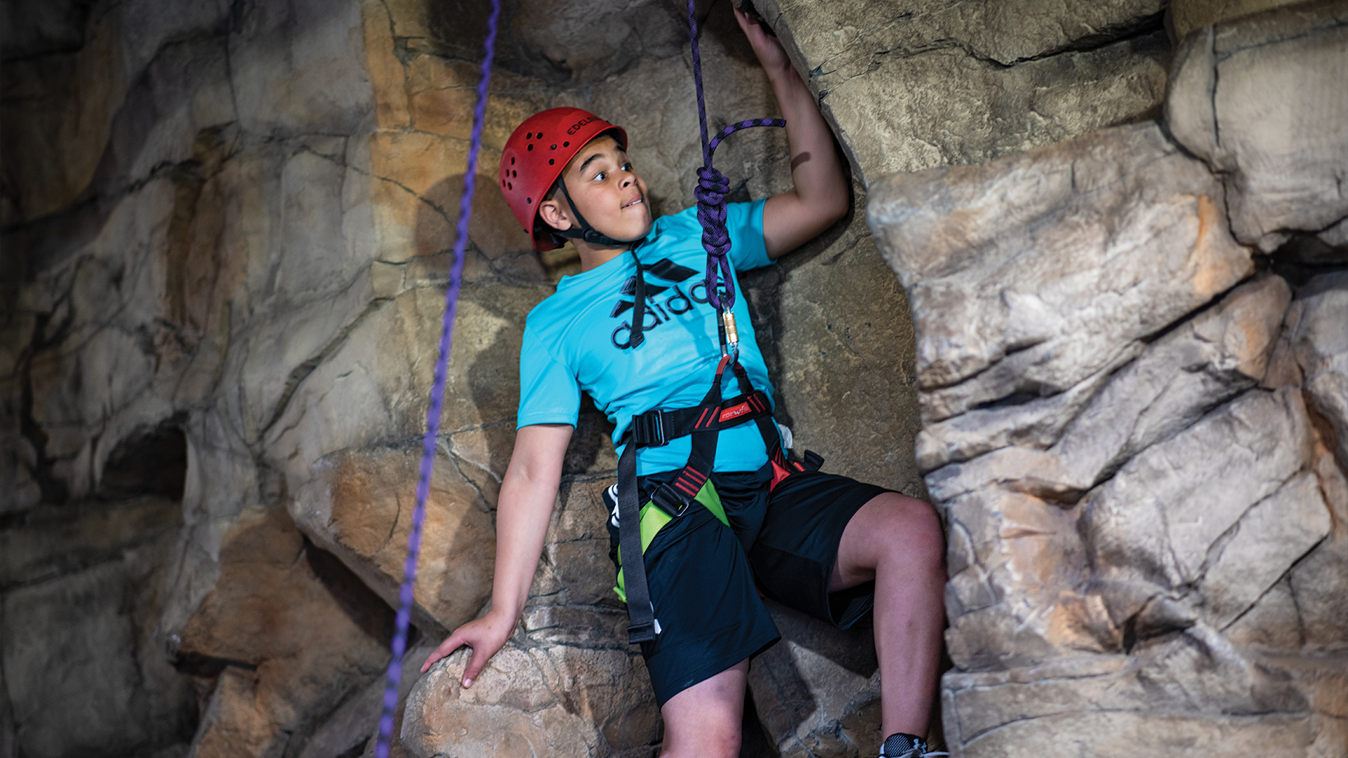 A photo of a young boy in a helmet and harness hooked into a rope climbing up an indoor rock wall