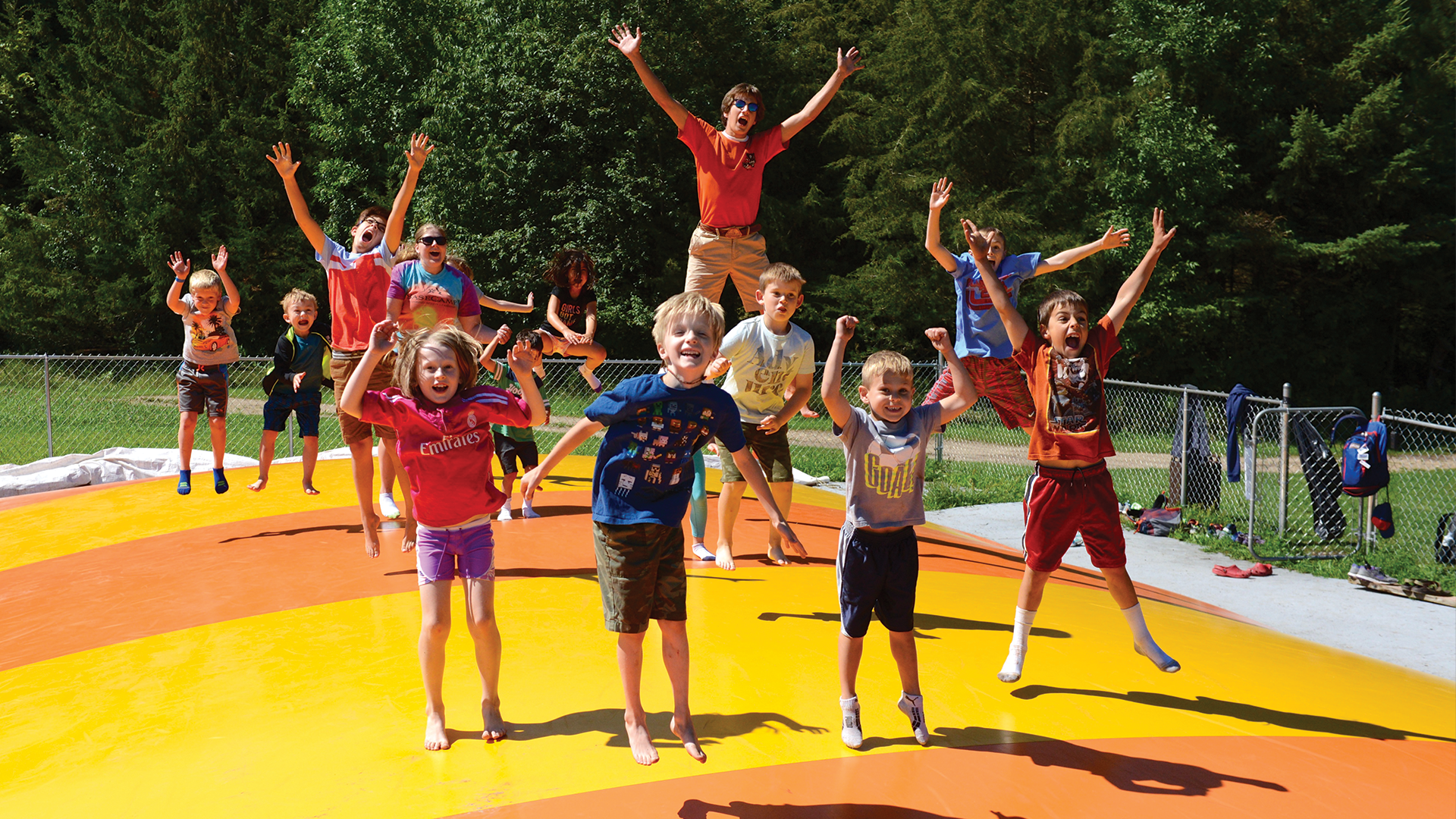 A group of 12 elementary-aged youth bouncing on the bounce pillow with some staff members. All appear to be excited and having a good time.