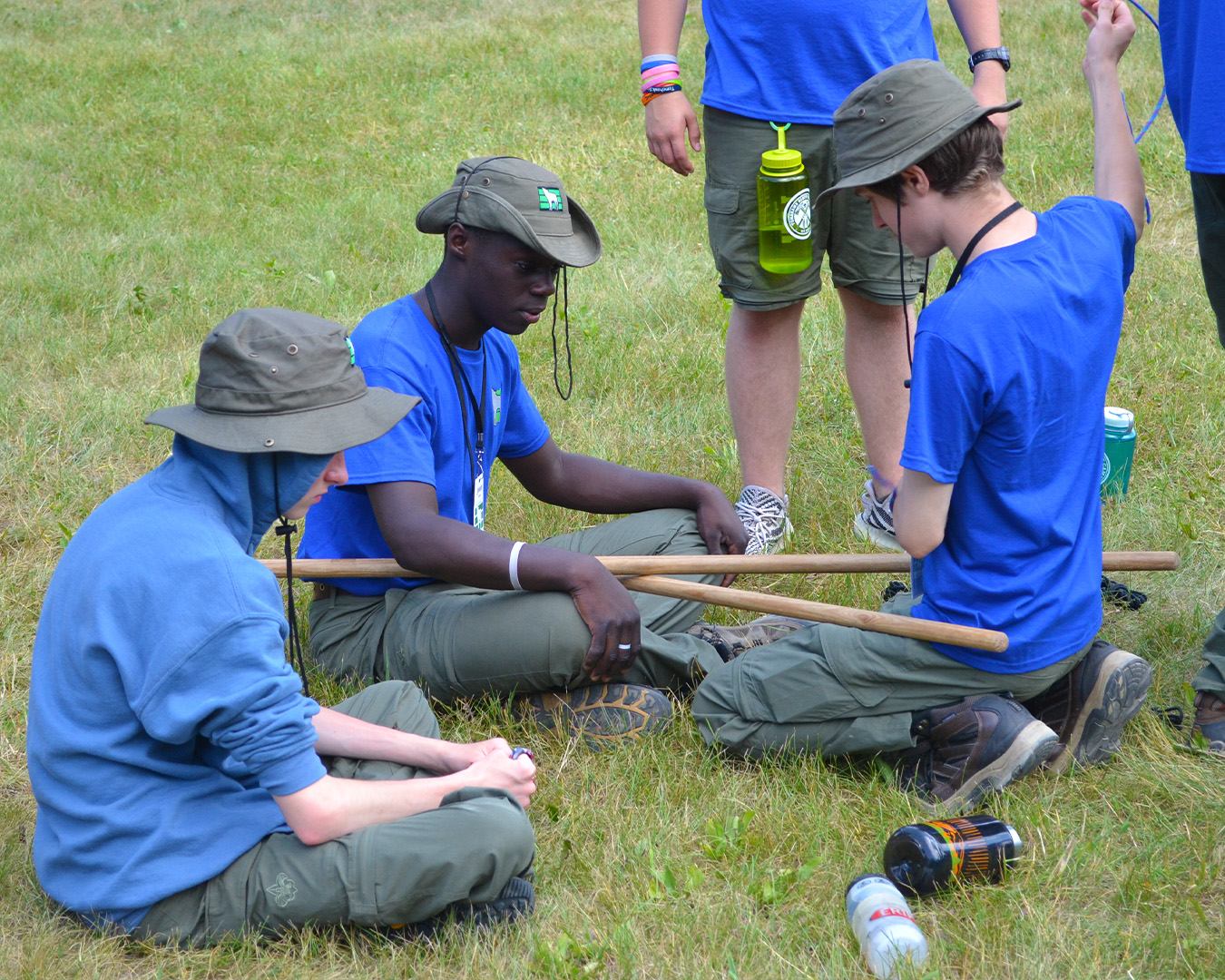 A group of Scouts sitting in the grass work on lashings while wearing the grey wolf shirt (a blue t-shirt with the grey wolf logo on the chest)