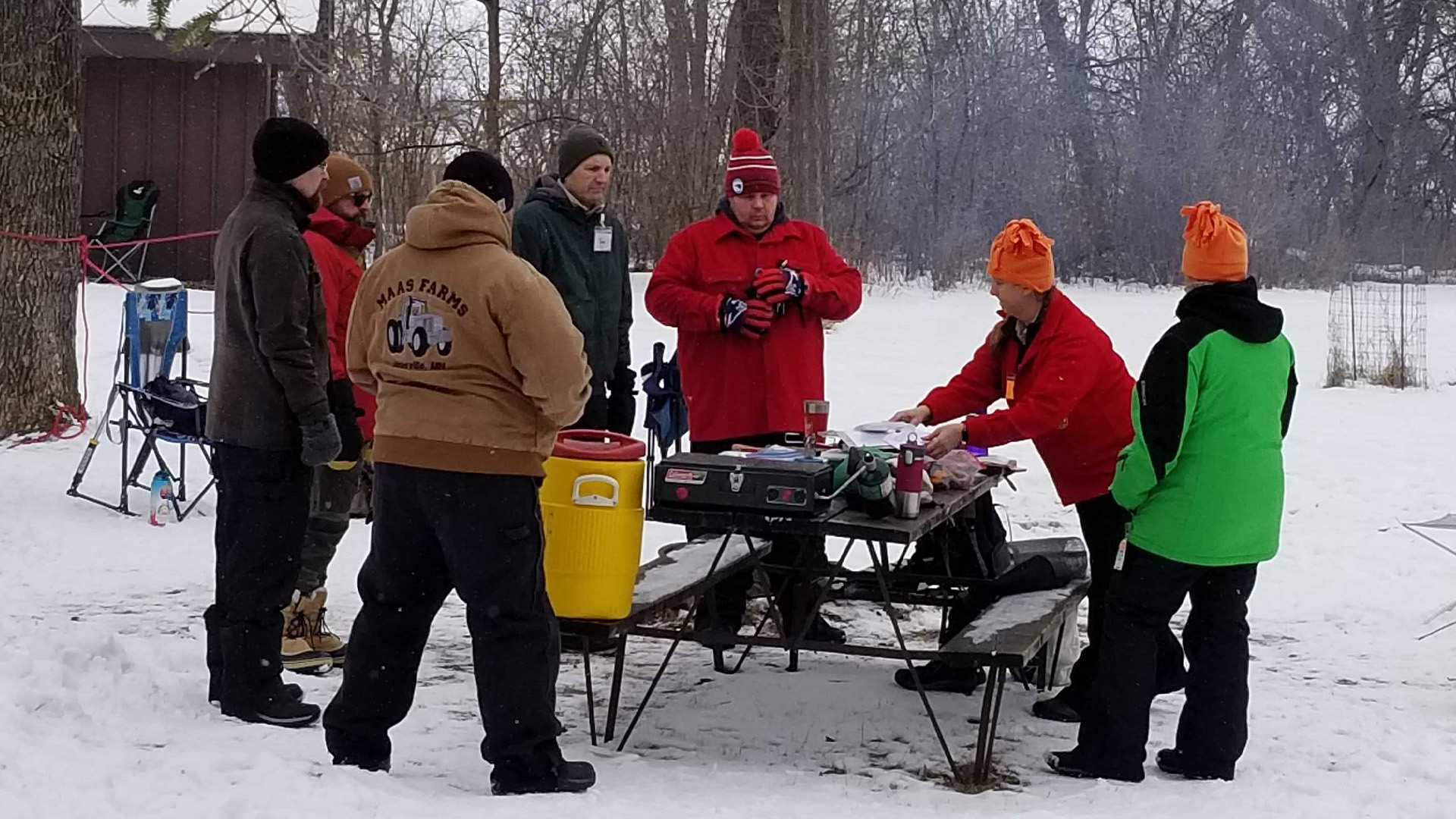 A group of adult leaders are all circled around a picnic table where they are cooking in the winter.