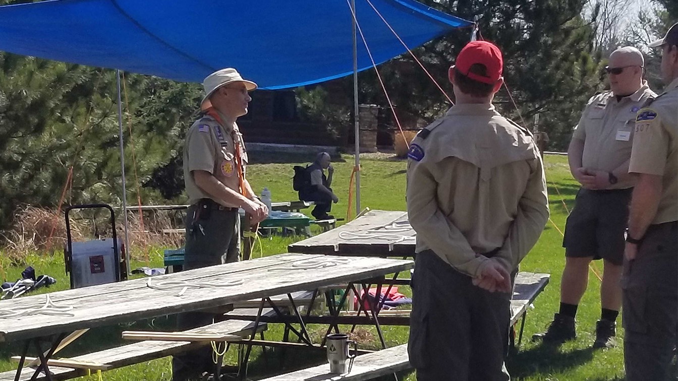 A course leader is teaching a group of adult leaders how to tie some knots using the EDGE method on picnic tables in the Spring.