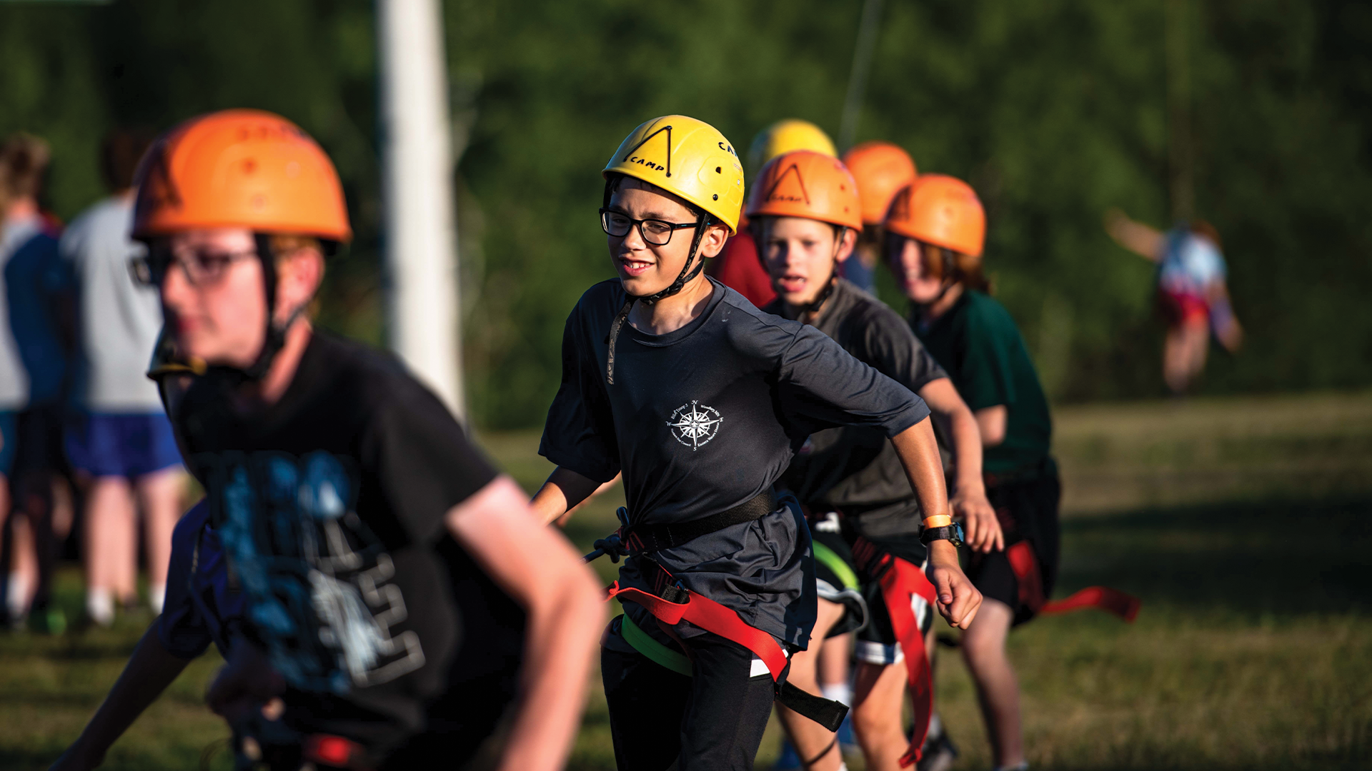A group of middle-school aged Scouts wearing climbing gear, hooked into a rope and running toward the camera. In the background, a Scout who is hooked into the same rope is beginning to float off of the ground.