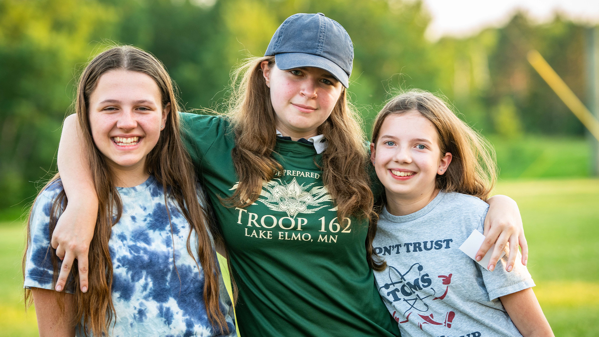 Three girls are smiling with their arms around each other while in the outdoors. The girl in the middle is wearing a shirt that say "Be Prepared - Troop 162 of Lake Elmo, MN".