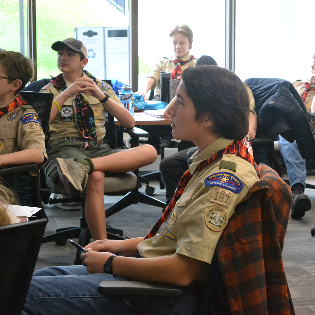 A large group of Scouts in uniform are sitting at desks in a classroom while an adult leader is pointing to a lesson slide outside of the frame