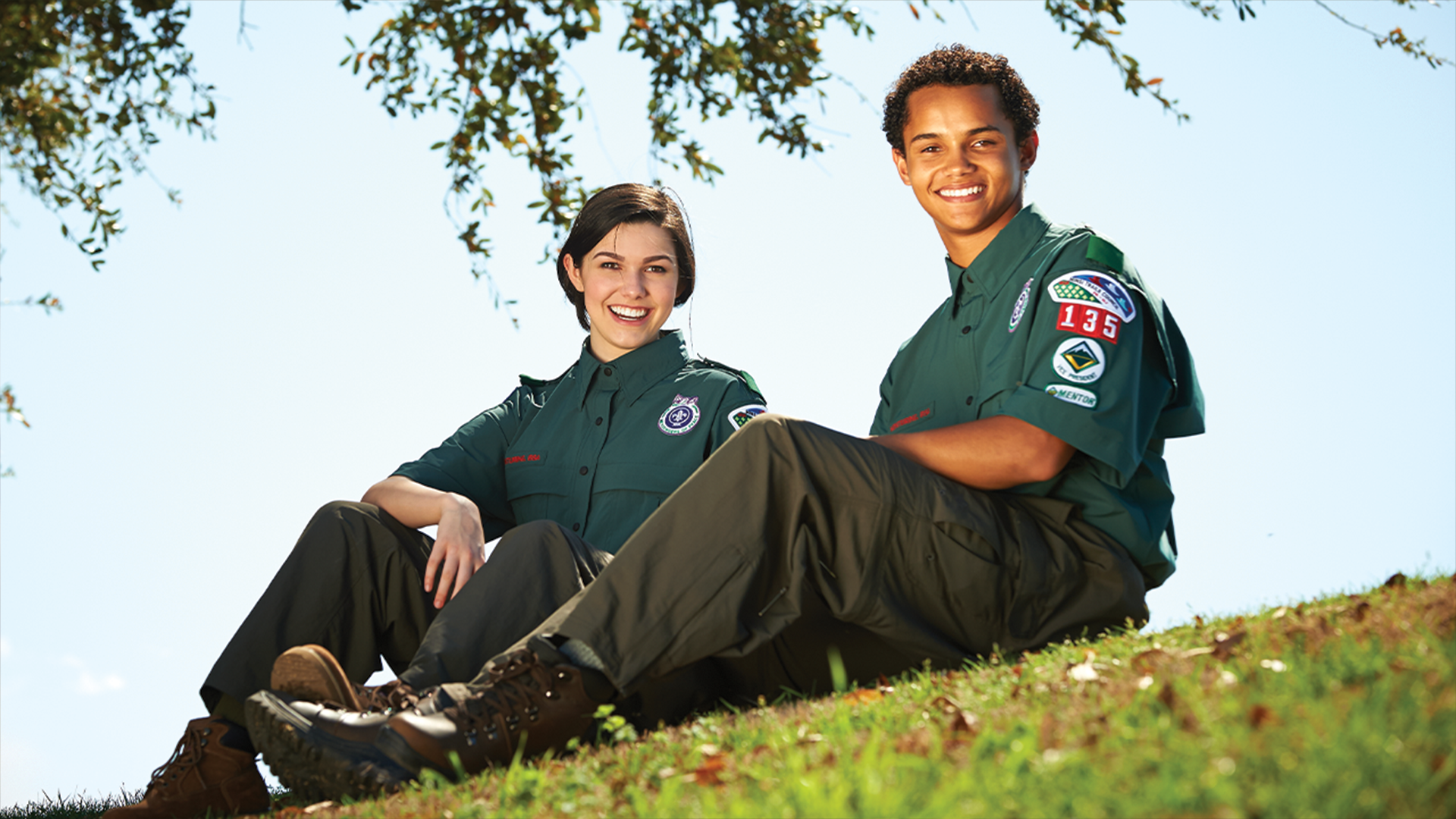 Two young adults wearing Venturing uniforms, one female and one male, are smiling at the camera while sitting on a hill 