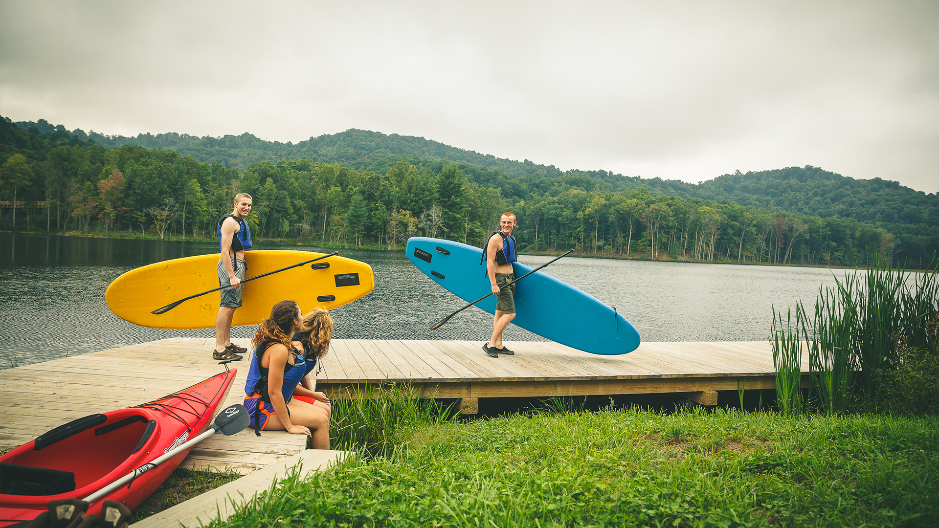 Two older male Venturing Scouts wearing lifejackets carry paddleboards and paddles on a dock on the shore of a lake. Two more older female Venturing Scouts sit on the dock after getting back from kayaking.