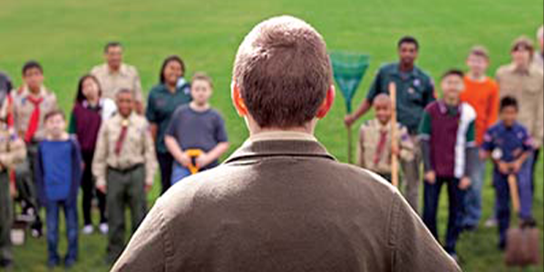 Picture of adult leader looking out on a group of people. All branches of Scouting and several community members are shown in the crowd.